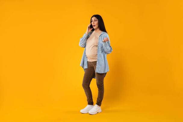 Young emotional pregnant woman talking on cellphone and gesturing, having phone conversation with lover or doctor while standing over yellow studio background, full length shot, free space - 写真・画像