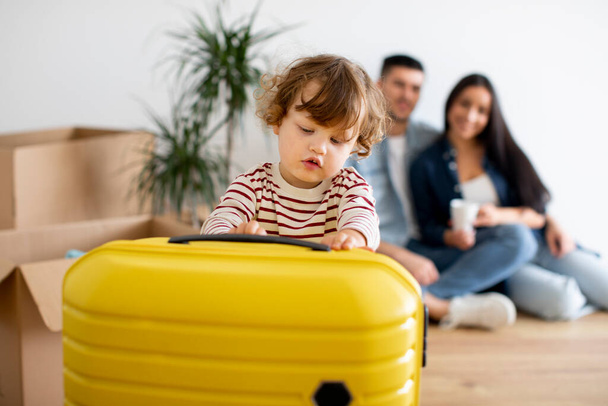Netter kleiner Junge, der mit dem großen gelben Koffer spielt, während er mit den Eltern nach Hause zieht, glückliche junge Familie von drei, die umziehen, sich im Wohnzimmer in ihrer neuen Wohnung ausruhen, selektiver Fokus - Foto, Bild
