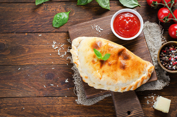 Pyszna pizzeria Calzone, tradycyjna włoska pizza z pomidorami i świeża bazylia na drewnianym rustykalnym tle - Zdjęcie, obraz