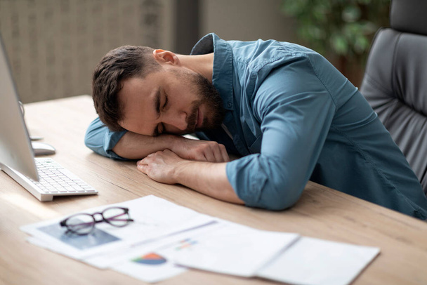 Exhausted young man sleeping on desk in office, next to laptop and documents, millennial male employee tired of overworking, workaholic guy suffering from chronic fatigue at workplace, closeup - Photo, Image