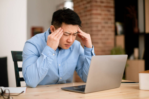 Stress and healthcare concept. Stressed asian middle aged businessman suffering from painful headache while working on laptop at home. Man having migraine symptoms at workplace - Fotografie, Obrázek