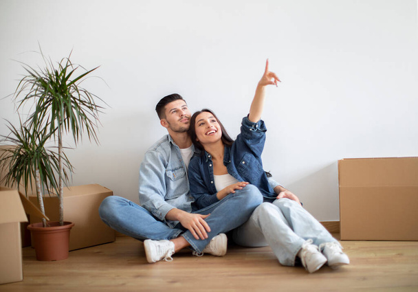 Snový mladý pár plánuje design interiéru po přestěhování domů, přemýšlí o dekoracích a nábytku ve svém novém domě, usmívá se evropští manželé sedí na podlaze a ukazuje na kopírovací prostor - Fotografie, Obrázek