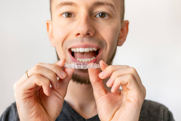 Young happy man with beard and white healthy smile is standing and demonstrating clear aligner for orthodontic correction of bite. Isolated background and close-up - Photo, Image