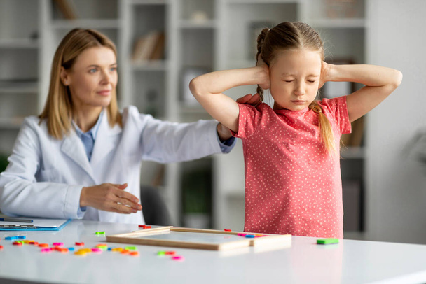 Professional Psychotherapist Lady Trying To Talk To Difficult Kid, Grumpy Little Girl Covering Ears And Closing Eyes, Disobedient Female Child Ignoring Psychologist During Session, Closeup - Photo, image