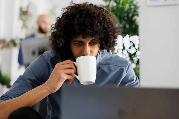 Arab employee managing seo optimization on laptop while drinking coffee in corporate office. Young professional holding tea cup and analyzing report analytics data in coworking space - Photo, Image