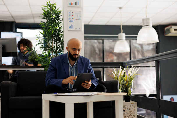 Arab businessman using tablet for digital work in business office. Zaměstnanec společnosti procházející sociální média během přestávky, zatímco sedí na gauči ve společném prostoru - Fotografie, Obrázek