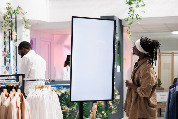 African american customer using smart board with empty screen to check information shoes collection in shopping mall. Man examining promotion on whiteboard in department store - Photo, Image
