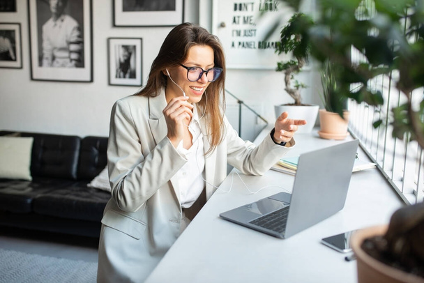 Pozitivní evropská žena manažerka ve sluchátkách a brýlích pracující na počítači, konzultace s klienty on-line pomocí PC, rozhovor s webkamerou, posezení v útulném prostoru - Fotografie, Obrázek