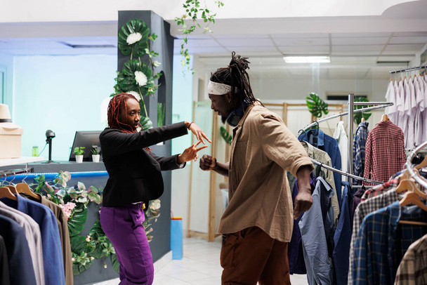 Αφρικάνικο ζευγάρι που απολαμβάνει τη μέρα του χορεύοντας και ακούγοντας μουσική στο εμπορικό κέντρο. Ξέγνοιαστοι άνδρες και γυναίκες φίλοι διασκεδάζουν επιλέγοντας ρούχα στο κατάστημα ρούχων - Φωτογραφία, εικόνα