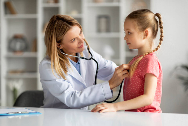 Female doctor checking lungs of little girl during medical checkup in clinic, friendly pediatrician woman using stethoscope to examine breathing and heartbeat of young patient, closeup shot - Photo, Image