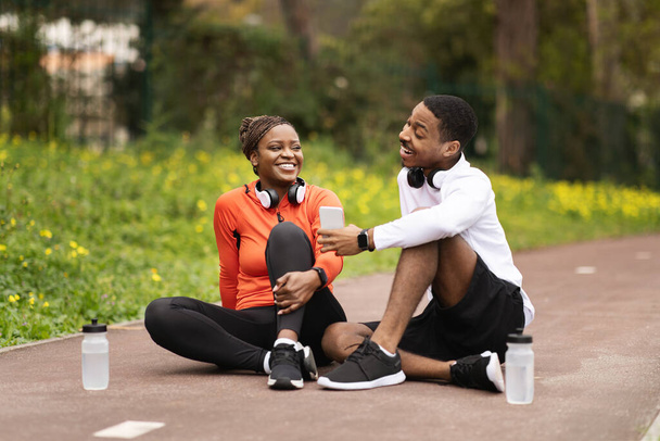 Happy young black couple sit on road with bottles of water, man recommended sport blog on phone in summer. Workout, exercise app, body care and relationship outdoor, advice - Photo, Image