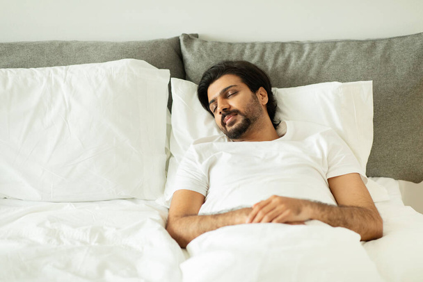 Millennial middle eastern guy sleeping, lies on white bed with empty space in bedroom interior. Sweet dream, loneliness, health care problems in relationship, ad and offer, top view - Foto, afbeelding