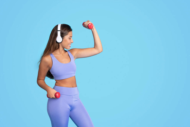 Happy young european slim woman athlete in sportswear and wireless headphones doing exercises with dumbbells, isolated on blue studio background. Sports, fit, body care and workout with music - Фото, зображення