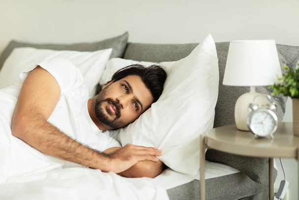 Sad serious middle eastern man woke up, lies on bed, looks at alarm clock in morning, suffer from depression in bedroom interior. Time to work, stress at home, ad and offer - Photo, Image