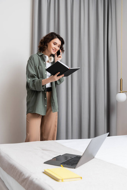 estilo de vida independiente, mujer feliz con auriculares inalámbricos mirando el cuaderno y hablando en el teléfono móvil cerca de cortinas grises, aplique de pared, ordenador portátil y bloc de notas en la cama cómoda en la habitación de hotel moderna - Foto, imagen