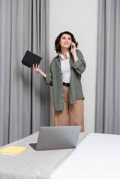 pleased woman with wavy brunette hair and wireless headphones holding notebook, looking away and talking on smartphone near grey curtains and laptop with notepad on comfortable bed in hotel room - Photo, Image