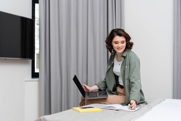 remote work, smiling woman with wavy hair writing in notebook and looking at camera while sitting near grey curtains, notepad and smartphone with blank screen on bed in modern hotel room - Photo, Image