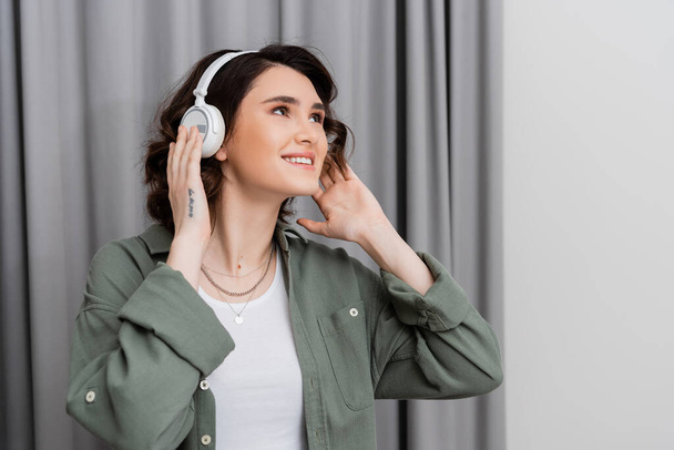 delighted young woman in casual clothes, with tattoo and with wavy brunette hair looking away while listening music in wireless headphones near grey curtains in modern hotel room, weekend getaway - Foto, afbeelding