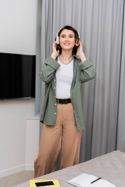cheerful and tattooed woman in casual clothes adjusting wireless headphones and looking away while listening podcast near grey curtains, lcd tv, notebooks, pen and smartphone with blank screen in hotel room - Photo, Image