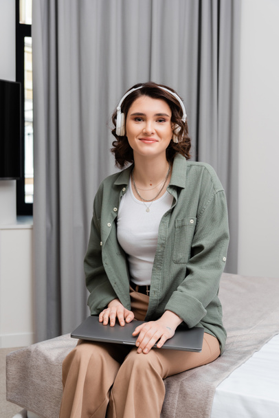 heureuse jeune femme aux cheveux bruns ondulés, en vêtements décontractés assis sur le lit avec ordinateur portable, écoutant podcast dans casque sans fil près de rideaux gris pendant l'éducation en ligne dans la chambre d'hôtel - Photo, image