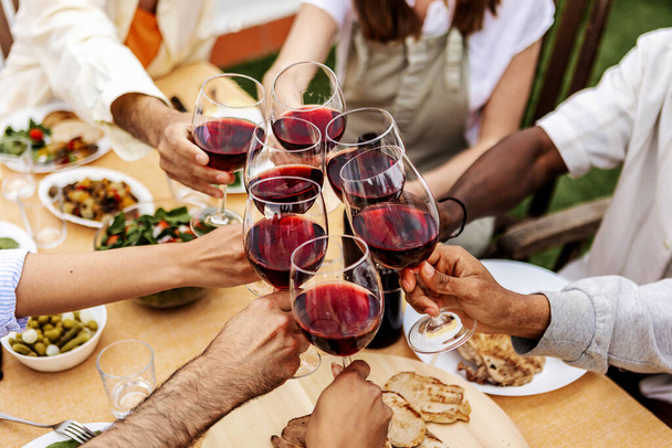 Grupa młodych ludzi wznosi toast za czerwone wino na przyjęciu na dachu. Wielorasowy przyjaciele świętujący na kolacji bbq w ogrodzie. - Zdjęcie, obraz