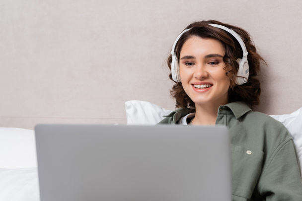 young and cheerful woman with wavy brunette hair watching film on blurred laptop while sitting in wireless headphones near grey wall and white pillows in modern hotel room - Photo, Image