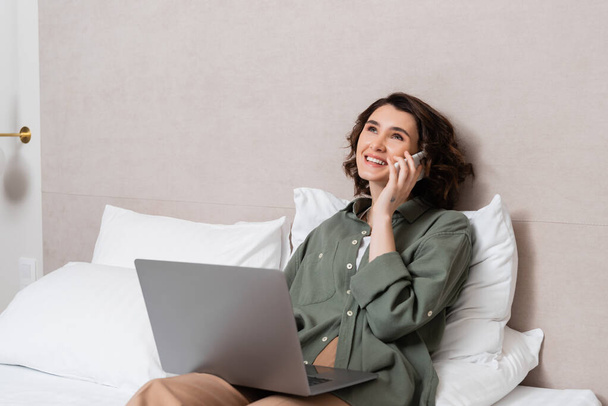 smiling woman with wavy brunette hair wearing casual clothes, holding laptop and talking on mobile phone while sitting on bed near white pillows in cozy hotel room, work-life integration - Photo, Image