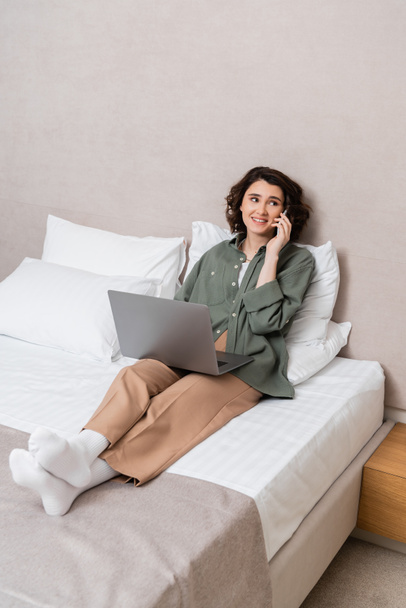 full length of young and happy woman in casual clothes, with wavy brunette hair looking away while sitting on bed with laptop and talking on mobile phone near white pillows in hotel suite - Photo, Image