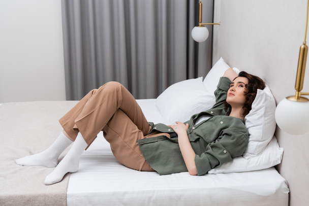 pełna długość marzycielska i beztroska kobieta z falującymi brunetkami układającymi się na łóżku w luźnych ubraniach w pobliżu białych poduszek, kinkietów i szarych zasłon w przytulnej atmosferze nowoczesnego pokoju hotelowego - Zdjęcie, obraz
