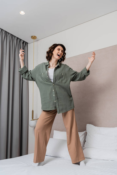 Dalgalı esmer saçlı genç bir kadın, günlük kıyafetler giyiyor, beyaz yastıkların yanında dans ediyor ve şarkı söylüyor, duvar lambası ve rahat otel odasındaki gri perdeler - Fotoğraf, Görsel