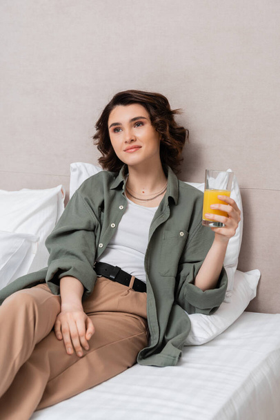zachwycona i marzycielska kobieta z falującymi włosami brunetki trzymająca szklankę świeżego soku pomarańczowego siedząc na łóżku w pobliżu białych poduszek i szarej ściany w nowoczesnym apartamencie hotelowym, wypoczynku i podróży - Zdjęcie, obraz