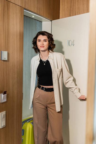 delighted woman in stylish clothes, white shirt and beige pants, with wavy brunette hair, holding yellow suitcase and opening door of modern hotel room with secure entry and keyboard access - Photo, Image