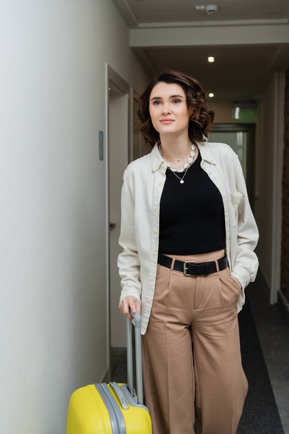 young woman with wavy brunette hair, in black crop top, white shirt and beige pants walking with hand in pocket and yellow suitcase along corridor in contemporary hotel  - Fotoğraf, Görsel