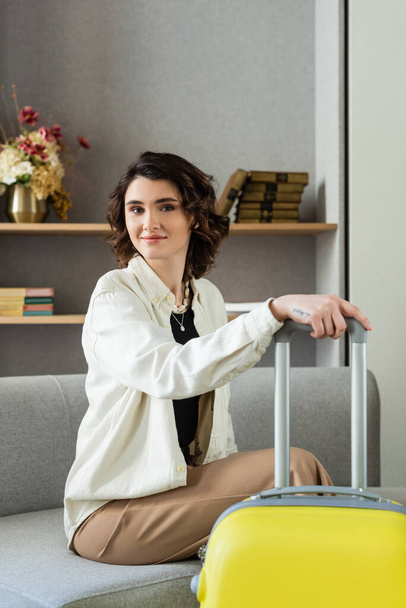 carefree tattooed woman with wavy brunette hair, in stylish casual clothes looking at camera while sitting on couch with yellow suitcase, near shelves with books and vase with flowers in hotel lobby - Photo, Image