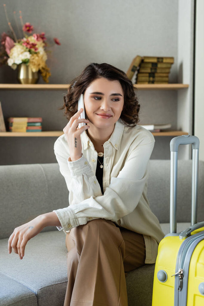 happy young woman with wavy brunette hair and tattoo sitting on couch and talking on mobile phone near yellow travel bag, books and vase with flowers on shelves in hotel lobby on blurred background - Photo, Image