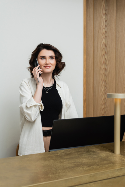 happy concierge with wavy brunette hair and tattoo wearing stylish clothes and talking on telephone at reception desk near computer monitor and lamp in lobby of modern hotel, customer service - Photo, Image