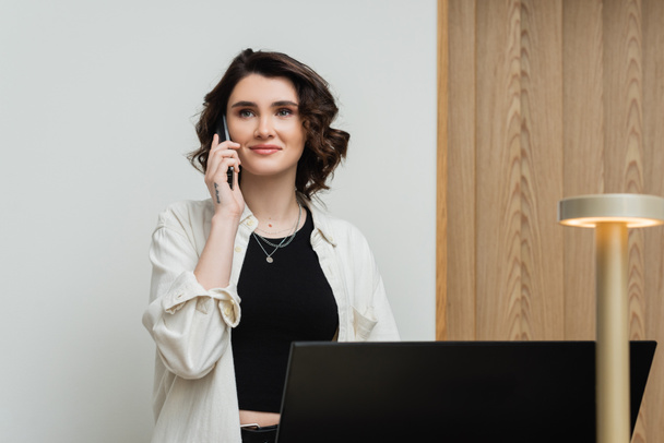 pleased young woman in stylish casual clothes, with wavy brunette hair and tattoo talking on telephone while working as receptionist at front desk near computer monitor and lamp in hotel lobby - Photo, Image