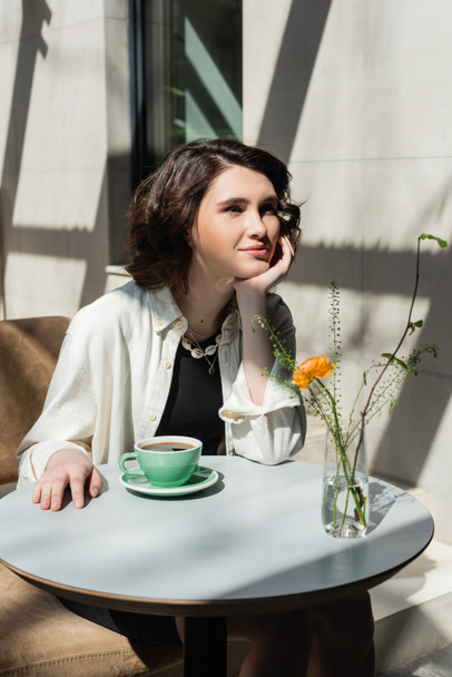 glimlachende vrouw zitten aan witte ronde tafel in de buurt van kopje zwarte aromatische koffie, schotel, glazen vaas met gele roos en groene planten op zomerterras van het hotel cafe in de ochtend zonlicht - Foto, afbeelding