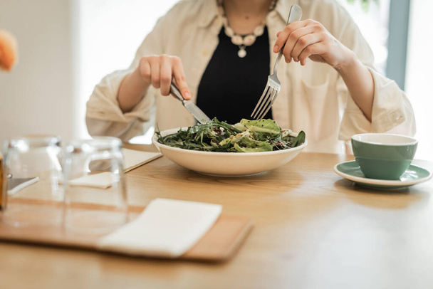 vista parcial de la mujer joven con tenedor y cuchillo comiendo ensalada de verduras frescas cerca de la taza de café aromático, platillo, vasos y servilletas en primer plano borroso en la mesa de la cafetería del vestíbulo en el hotel moderno  - Foto, Imagen
