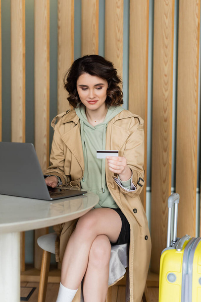 appealing young woman in stylish beige trench coat and with wavy brunette hair sitting at table with laptop and holding credit card during internet shopping in lobby of contemporary hotel - Photo, Image