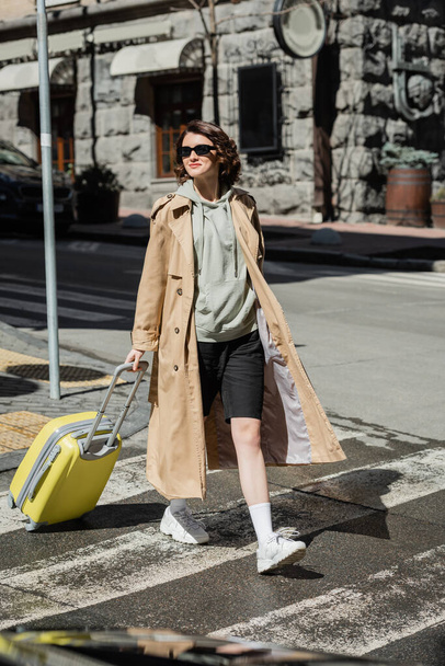 full length of self-assured and independent woman in beige trench coat, grey hoodie and black shorts holding yellow suitcase while crossing road in European city, urban lifestyle - Photo, Image
