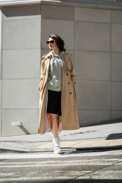 full length of trendy woman with wavy brunette hair crossing road in beige trench coat, grey hoodie, black shorts, white sneakers and dark sunglasses near modern building in city - Photo, Image