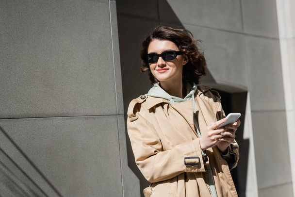 ευχαριστημένη και μοντέρνα γυναίκα με σκούρα γυαλιά ηλίου, κουκούλα και μπεζ παλτό μηνυμάτων σε smartphone και κοιτάζοντας μακριά, ενώ στέκεται κοντά σε γκρι κτίριο στην πόλη, αστικού τρόπου ζωής, φωτογραφία του δρόμου - Φωτογραφία, εικόνα