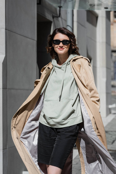 mujer elegante y despreocupada en gafas de sol oscuras, sudadera con capucha gris y gabardina beige posando en la calle en la ciudad europea moderna y mirando a la cámara, estilo de vida de viaje, moda urbana - Foto, Imagen
