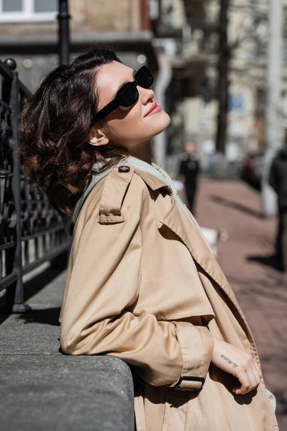 Seitenansicht einer jungen und unbeschwerten Frau mit welligem brünetten Haar und Tätowierung, in dunkler Sonnenbrille und beigem Trenchcoat, die sich in der Nähe des geschmiedeten Zauns in der europäischen Stadt entspannt, Straßenfotografie, urbane Mode - Foto, Bild
