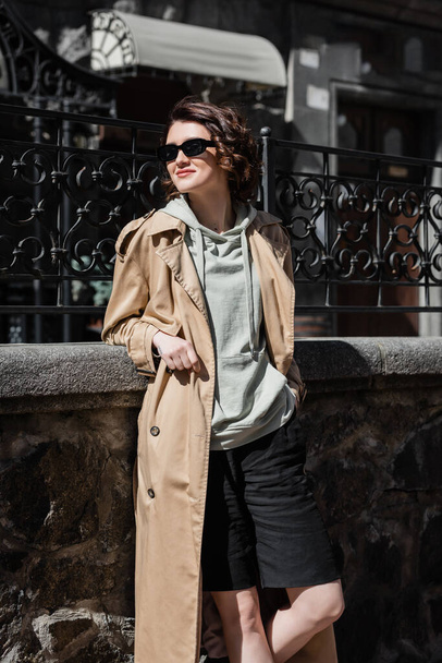 travel lifestyle, trendy and cheerful woman in grey hoodie, beige trench coat and stylish sunglasses standing with hand in pocket of black shorts and looking away near forged fence on city street - Photo, Image