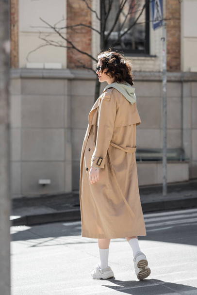 full length of young and trendy woman in grey hoodie, beige trench coat, white sneakers and dark sunglasses looking away and crossing in European city, street photography, urban fashion - Fotografie, Obrázek