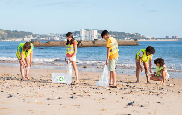 Радостные разнообразные подростки-волонтеры и женщины в жилетах собирают мусор в мешки для переработки на пляже, на открытом воздухе. Командная работа, забота об окружающей среде, экологическая природа, борьба с пластиком - Фото, изображение