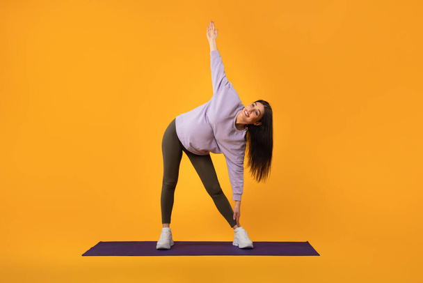 Prenatální fitness koncept. Mladá těhotná dáma zůstává aktivní, protahování nohou a rukou při cvičení na žlutém pozadí studia, plná délka, volný prostor - Fotografie, Obrázek