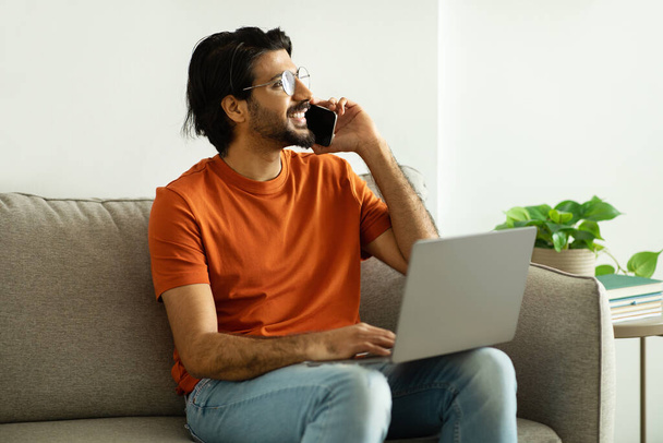 Positive millennial middle eastern man in glasses calls by phone, speaks with laptop, looks at free space, sits on sofa in living room interior. Communication, work remotely, device, ad and offer - Φωτογραφία, εικόνα